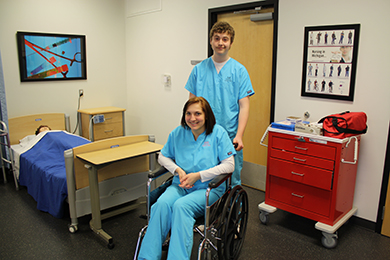A male Allied Health Technologies student pushes a female student in a wheelchair.