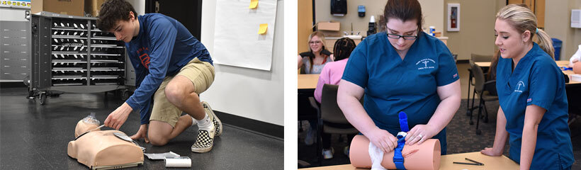 A male student practicing CPR on a dummy. Two female students standing over a table practicing packing a wound using a dummy leg.